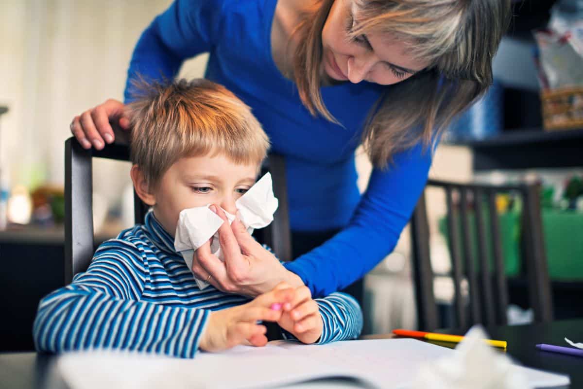 symptoms of pediatric allergies Mother leaning over child in kitchen chair as they hold a tissue up to a sitting child's nose