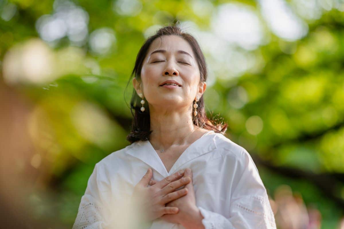 The effects of latera on a person with nasal obstruction Woman in a park. Upper body and head are in frame. Her hands are clasping her chest. Her head is tilted slightly back and her mouth is barely open. Her eyes are closed in a look of joyful contemplation. The background is very blurry but includes the shape of trees and people.