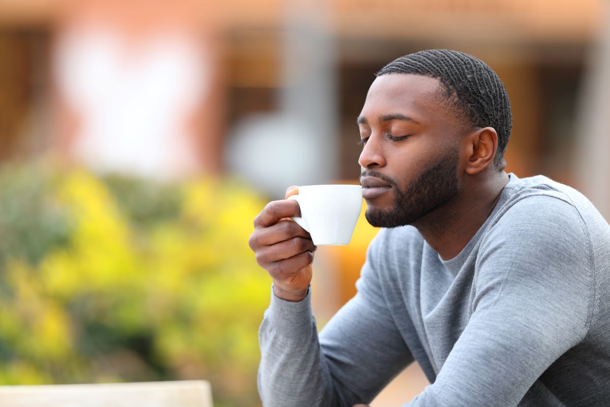 effects of loss of smell Man smelling a cup of coffee in an outdoor setting containing a bush of yellow flowers and a blurry storefront