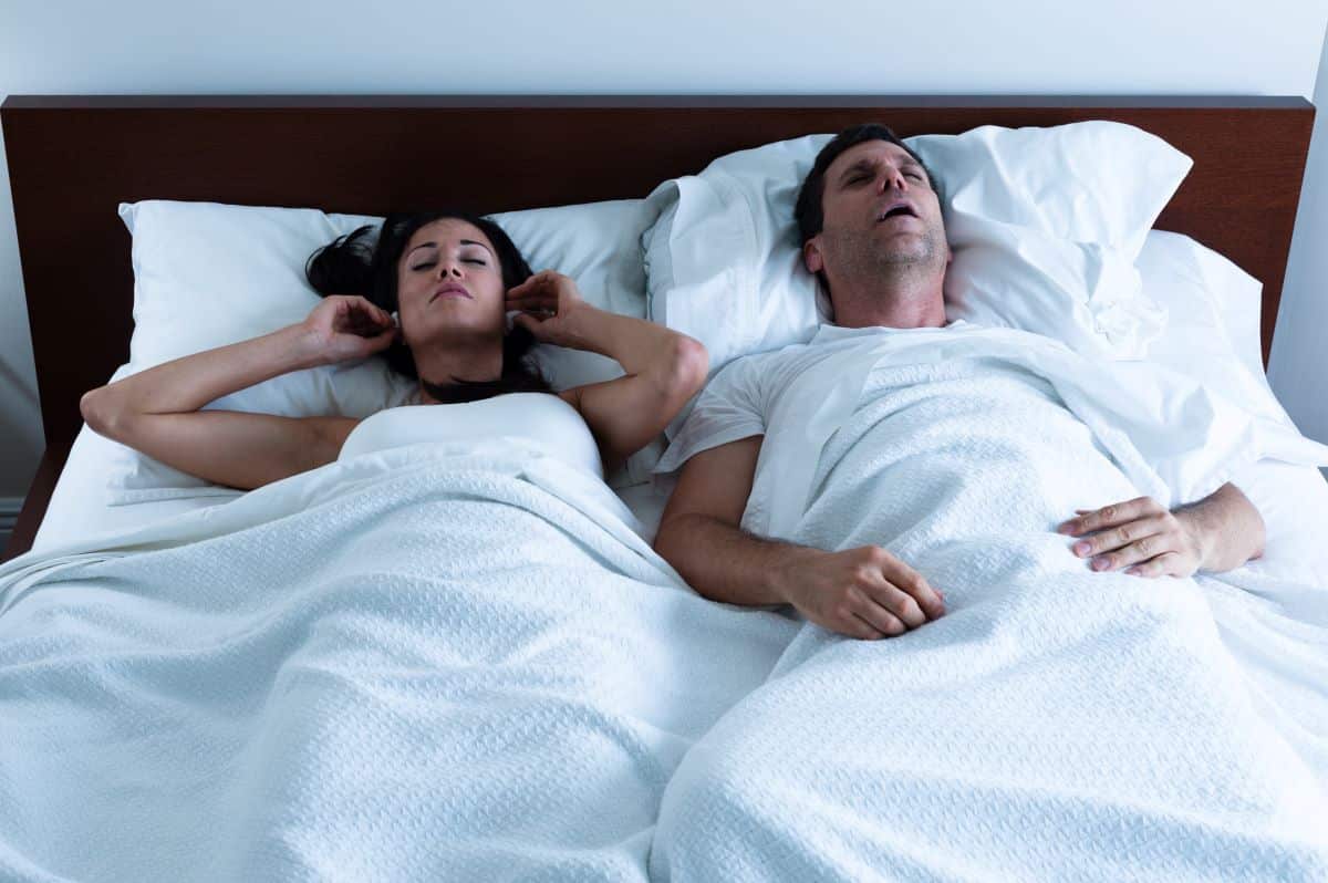 snoring-symptoms A heterosexual couple in bed. The man on the right has his mouth open as though snoring. The woman on the left is placing ear plugs into her ears.