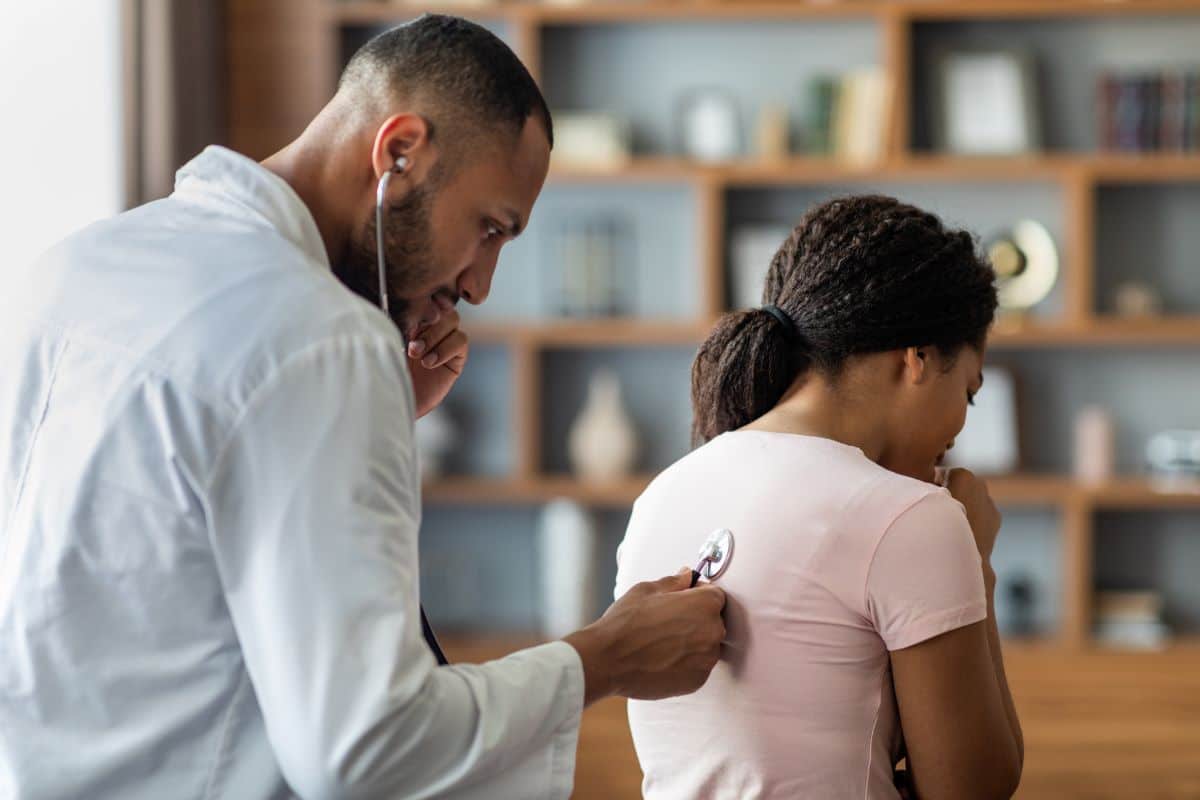 patient with chronic cough getting diagnosed Male doctor in an office setting holding a stethoscope to a female patients back to listen to their lungs