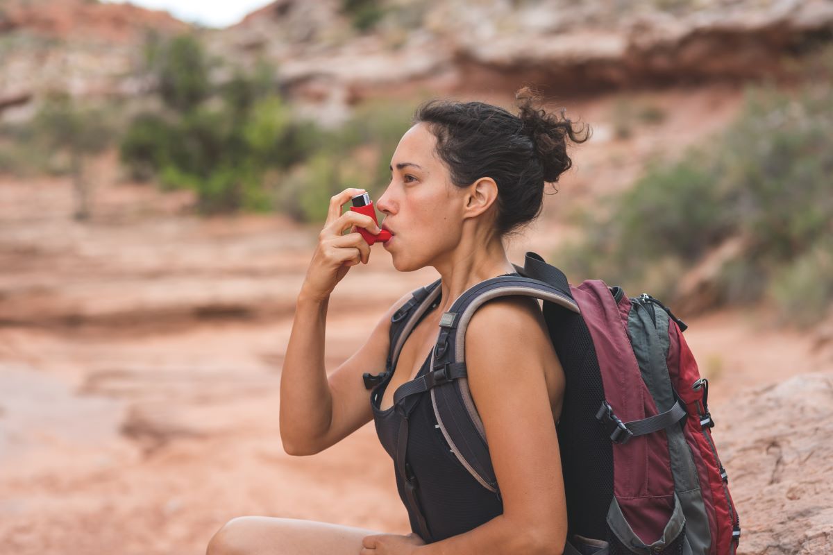 woman using asthma inhaler Woman with a backpack on in a desert setting is using a red inhaler.