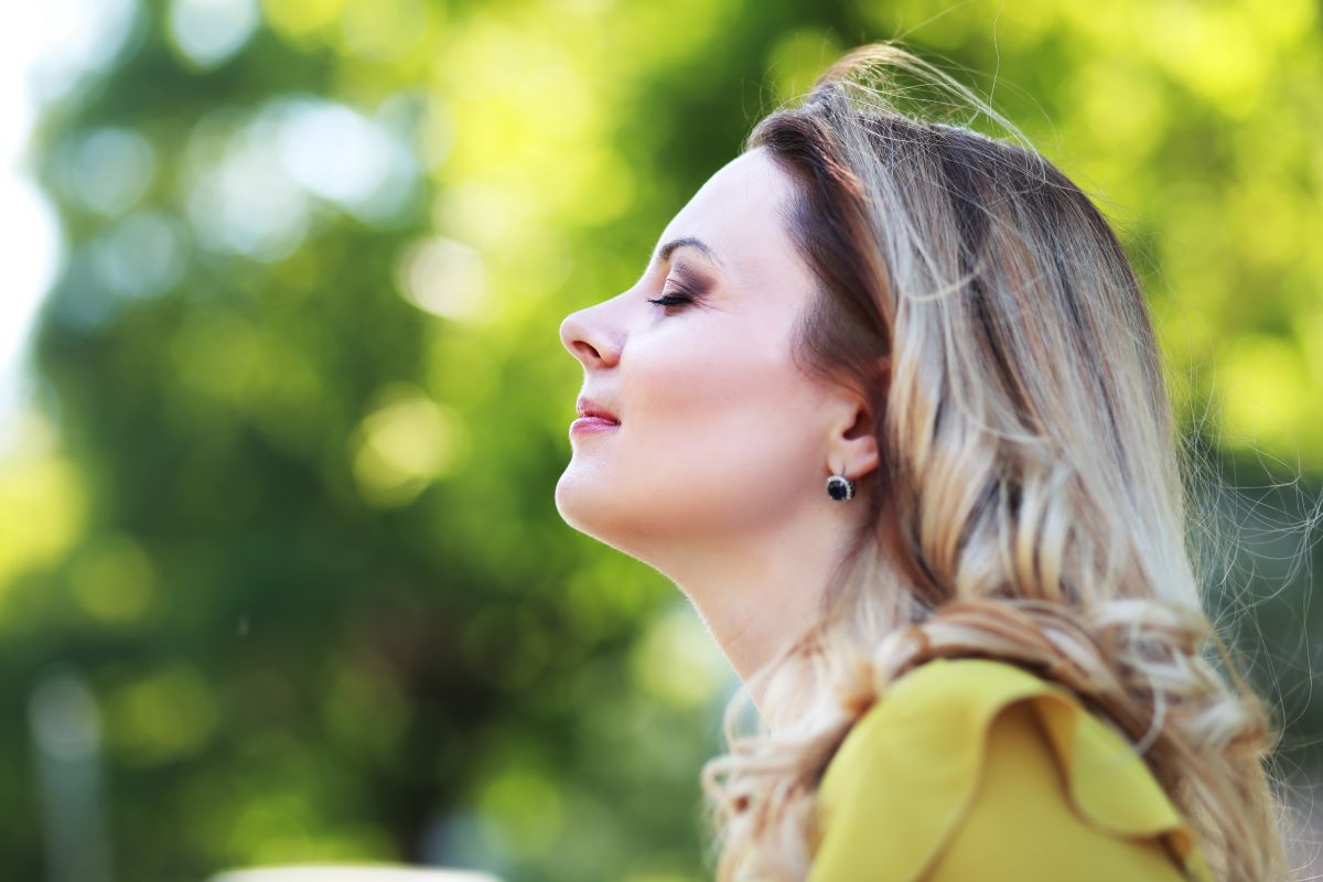 Rhinitis Sufferer Needing Propel Sinus Implant Woman in profile in an outdoor setting. She is facing the left side of the frame with her eyes closed and her chin tilted slightly up. She has a content expression. The background is heavily blurred tree canopy with enough light to indicate it is not a forest.