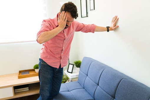 Balance Vertigo Issues Man in pink shirt in an office setting holding his head with one hand while propping himself up against a wall with his other arm outstretched.
